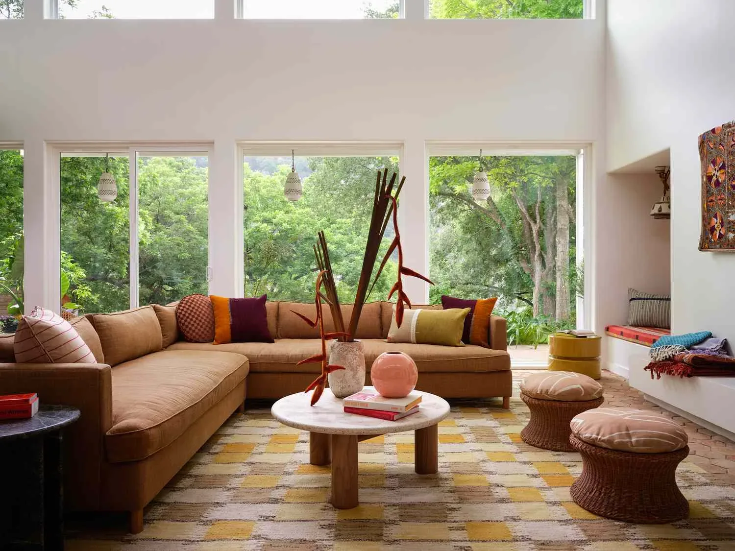 Warm Earth-Tone Living Room Designed by Decozie Designer Nicole W. Spacious living room with a caramel sectional sofa, earth-tone accent pillows, round wooden coffee table, patterned rug, and floor-to-ceiling windows overlooking lush greenery, designed by Decozie designer Nicole W.
