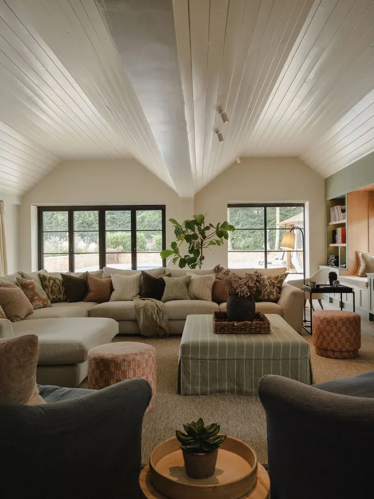 Cozy and Stylish Living Room with Vaulted Ceiling A photograph of a comfortable living room with a white vaulted ceiling and large windows. It features an L-shaped sofa, a striped ottoman, and a fiddle leaf fig plant. Two armchairs are in the foreground.