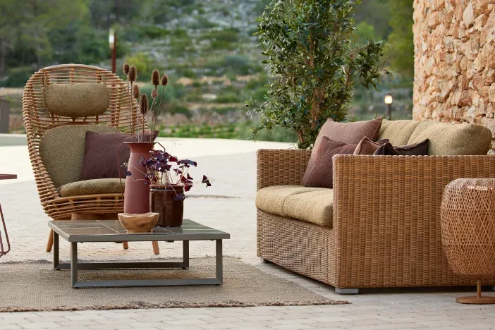 Cozie Outdoor Sanctuary: Natural Rattan Cocoon Chair and Sofa Curated outdoor living space on a patio, featuring a high-backed natural rattan cocoon chair and matching two-seater wicker sofa with olive green and brown cushions, on a natural jute rug. Background of a textured stone wall and Mediterranean hillside.