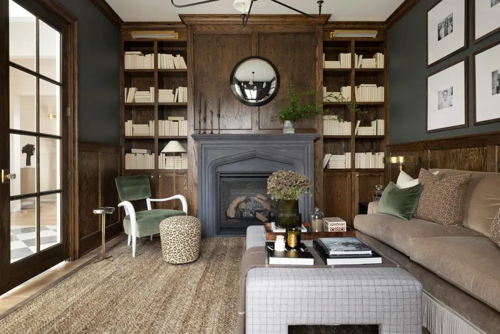 Moody Wood-Paneled Living Room with Built-In Library Designed by Decozie Designer Austin T. Sophisticated living room with dark wood paneling, built-in bookshelves, a black fireplace, neutral sofa with green accent pillows, and a cozy reading chair, designed by Decozie designer Austin T.