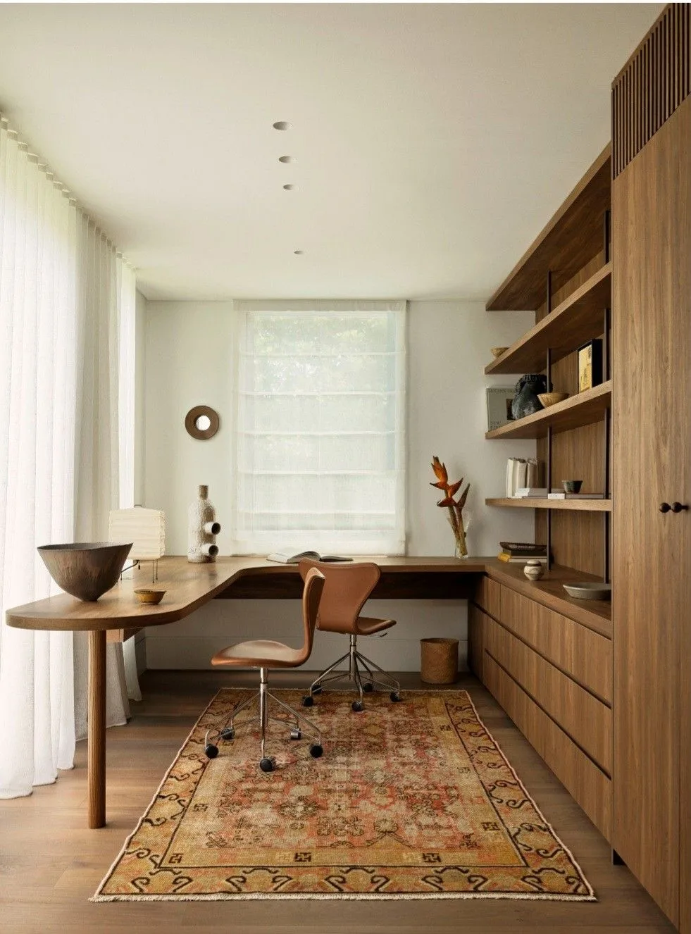 Modern minimalist home office with a wooden built-in desk and shelving, a leather swivel chair, sheer curtains filtering natural light, and a vintage patterned rug adding warmth to the space.Designed by Decozie with online interior design services.
