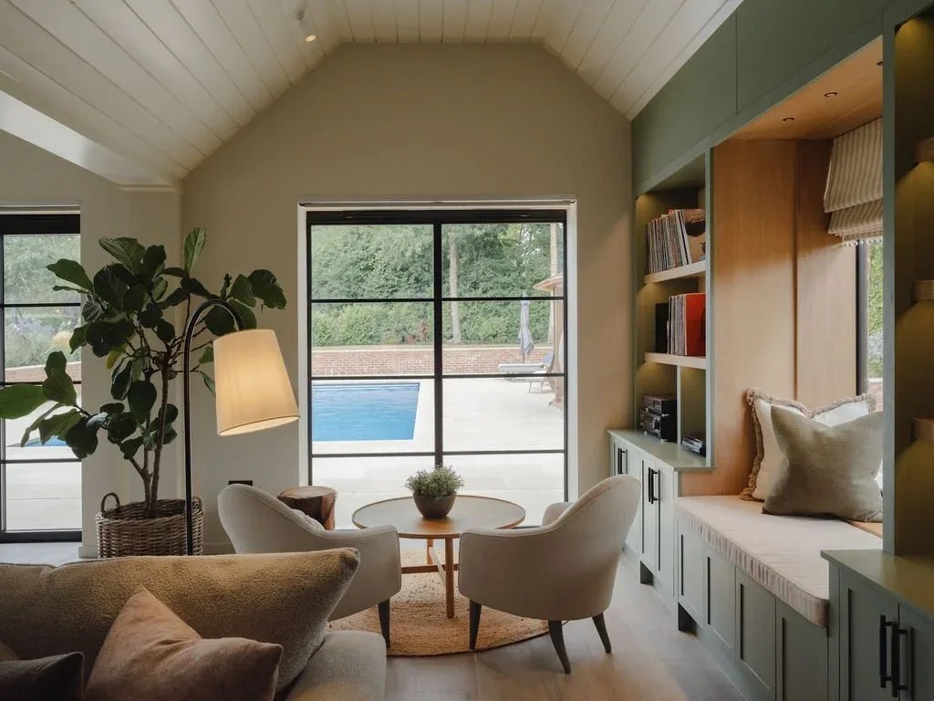 Modern Living Room Design with Neutral Tones and Pool View \"After\" Cozy modern living room with beige sofas, armchairs, floor lamp, built-in window seating, and indoor-outdoor pool view